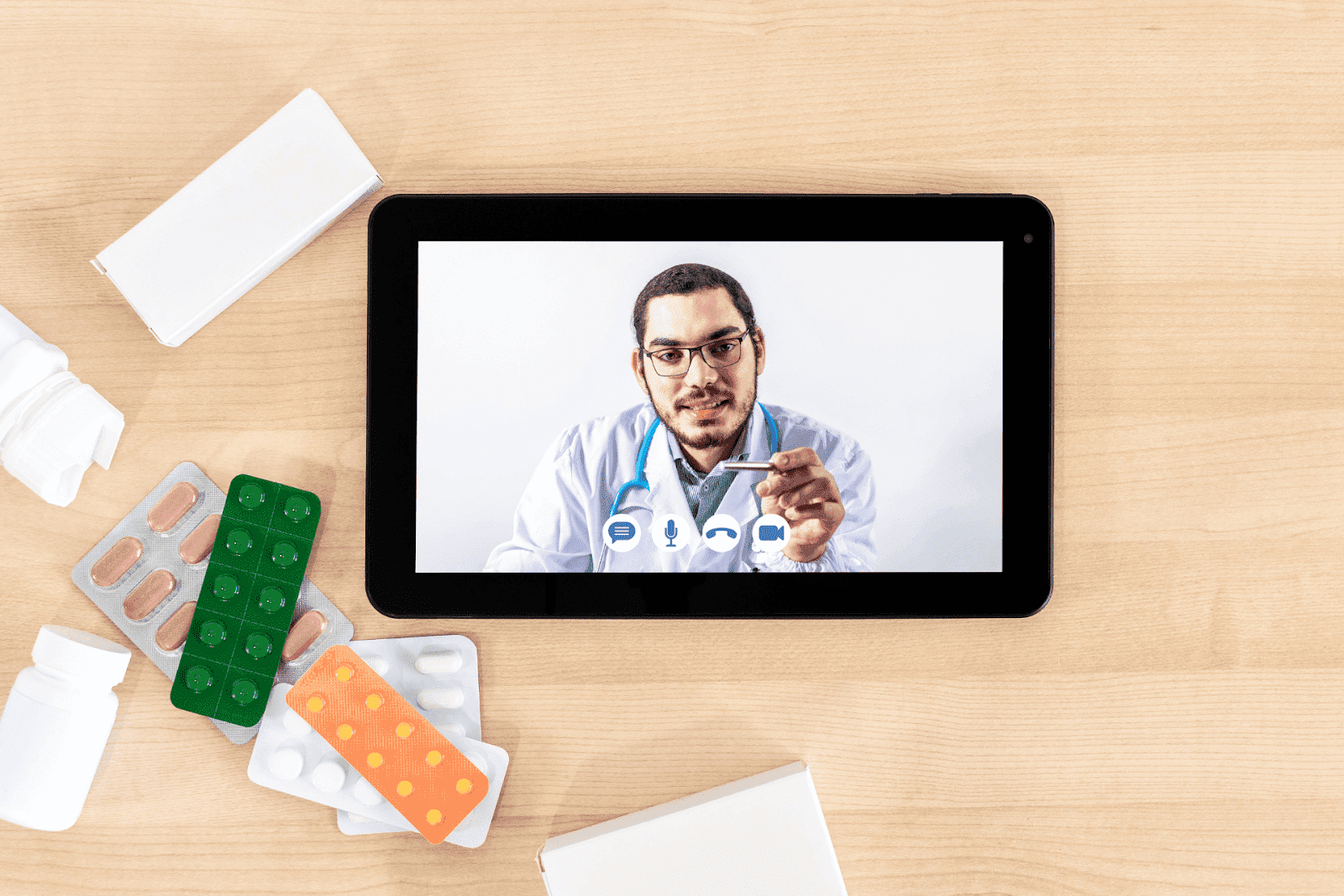 A doctor appears on a tablet screen during a virtual consultation, with various pill blister packs and bottles on the table.