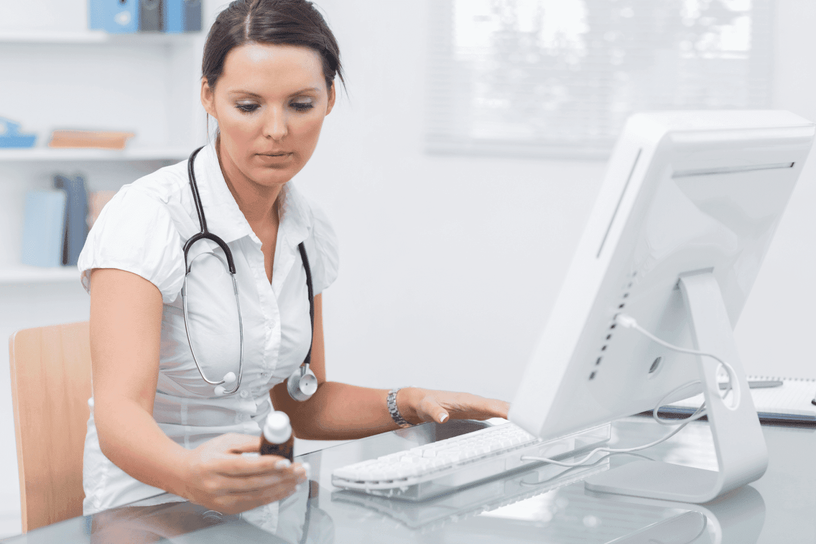 A doctor at a desk using a computer while holding a medicine bottle