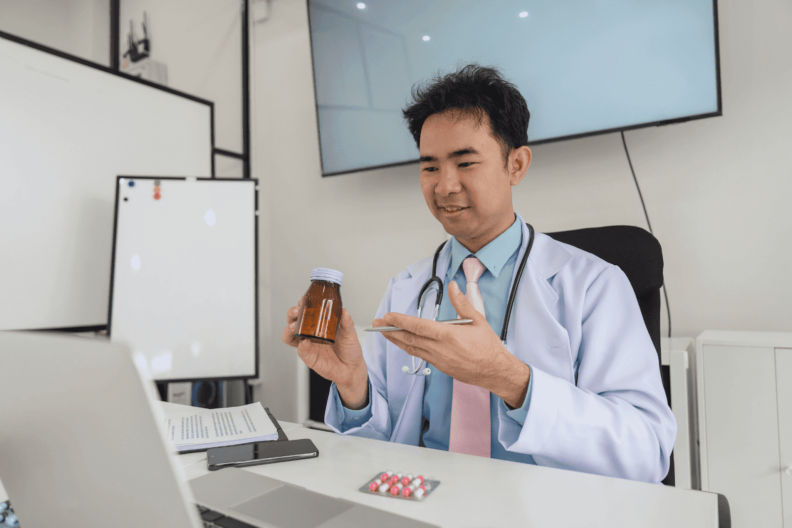 A doctor in a white coat and stethoscope shows a brown medicine bottle during a video consultation.