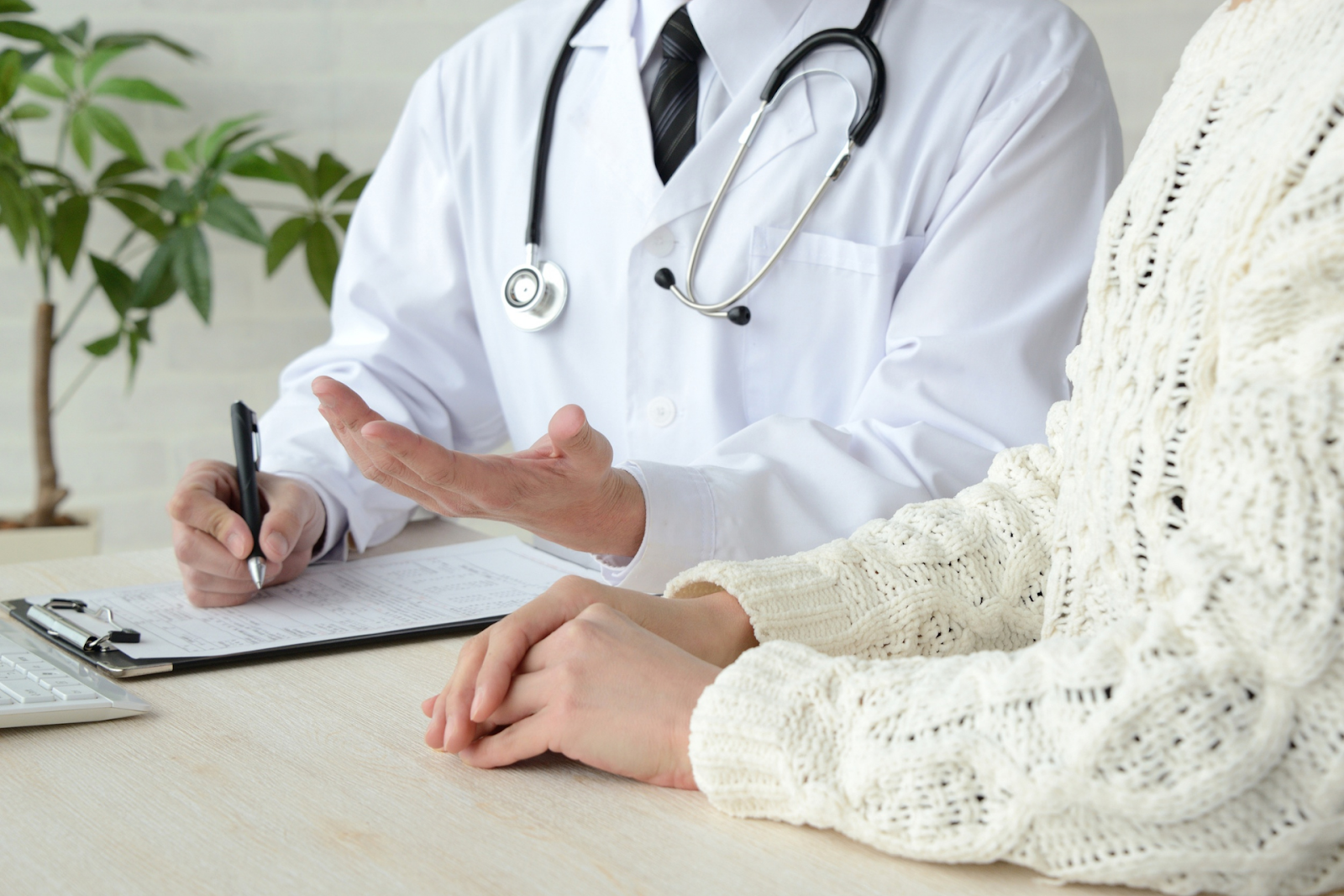 A doctor in a white coat is gesturing and writing on a clipboard while consulting a patient whose hands rest on the table.
