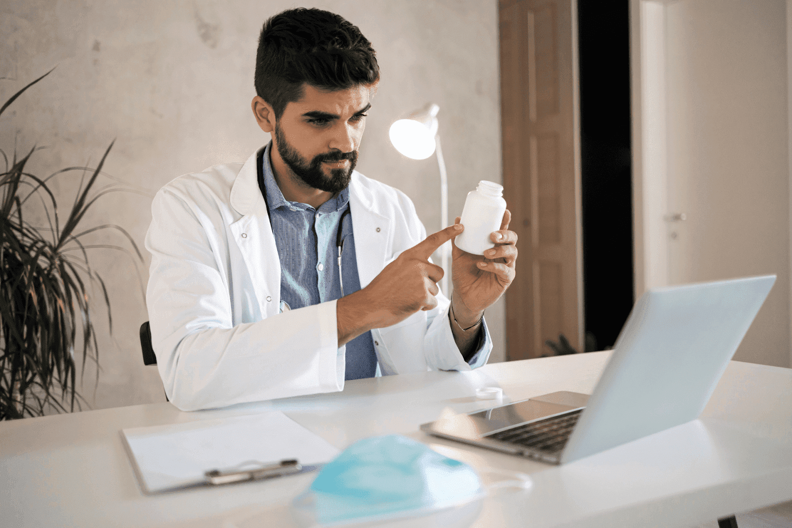 A doctor in a white coat points at a pill bottle while sitting at a desk with a laptop, clipboard, and face mask