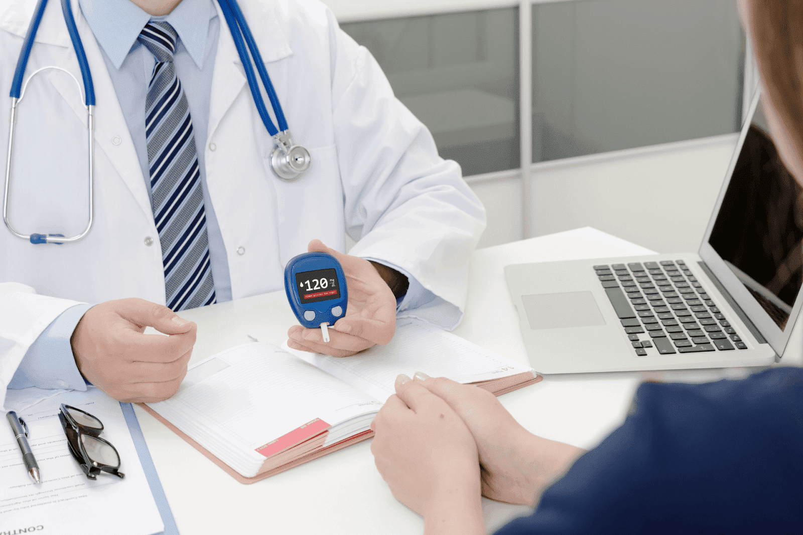 A doctor shows a patient a glucometer reading during a consultation at a desk with a laptop and medical notes
