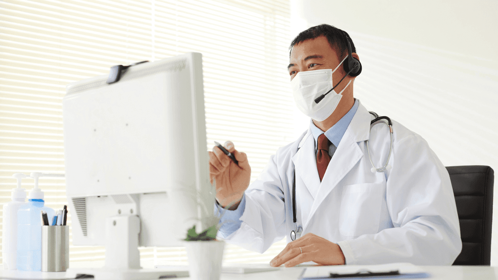 A doctor wearing a mask and headset sits at a desk using a computer for a virtual consultation.