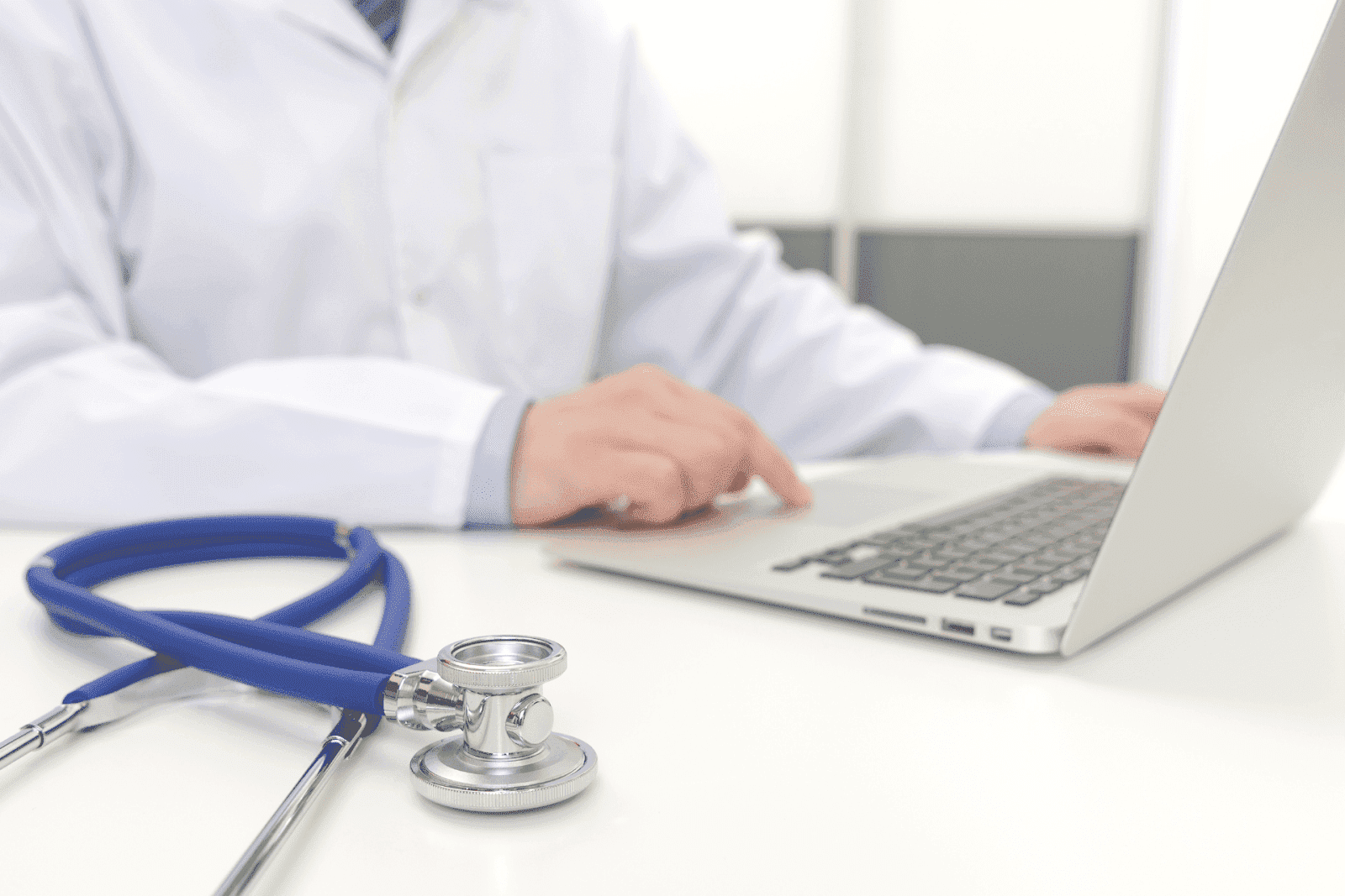 A doctor works on a laptop with a stethoscope lying on the desk nearby.