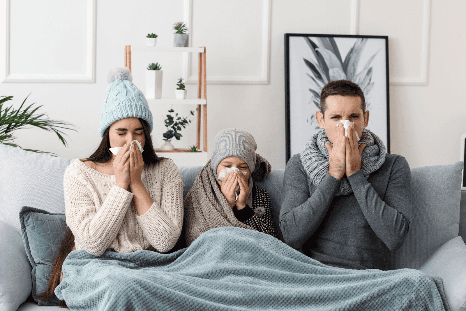 A family of three sitting on a couch under a blanket, all wearing winter clothing and holding tissues to their noses, appearing sick with flu-like symptoms, illustrating household flu spread.