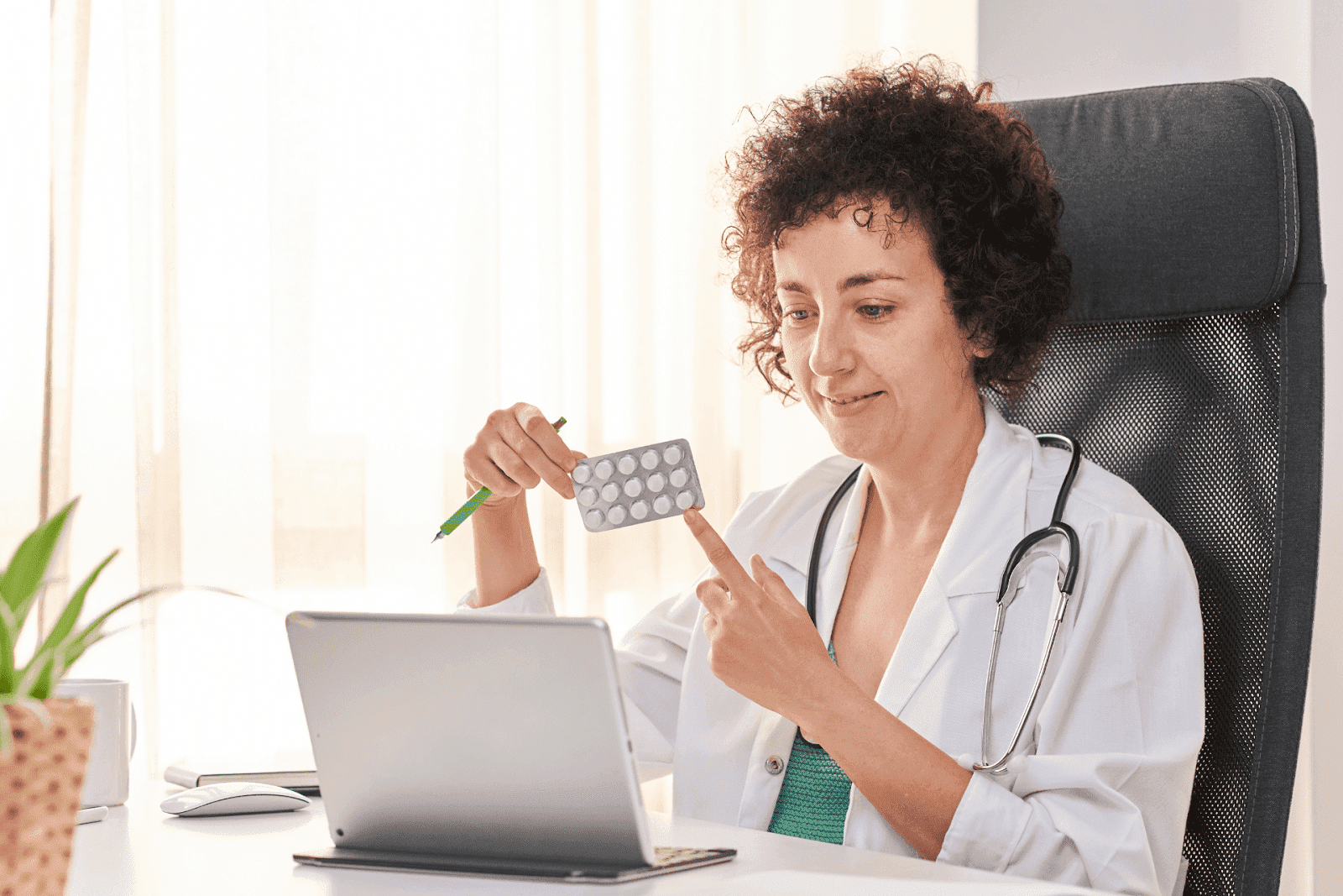 A female doctor in a white coat holds and points to a blister pack of pills while speaking during an online consultation.