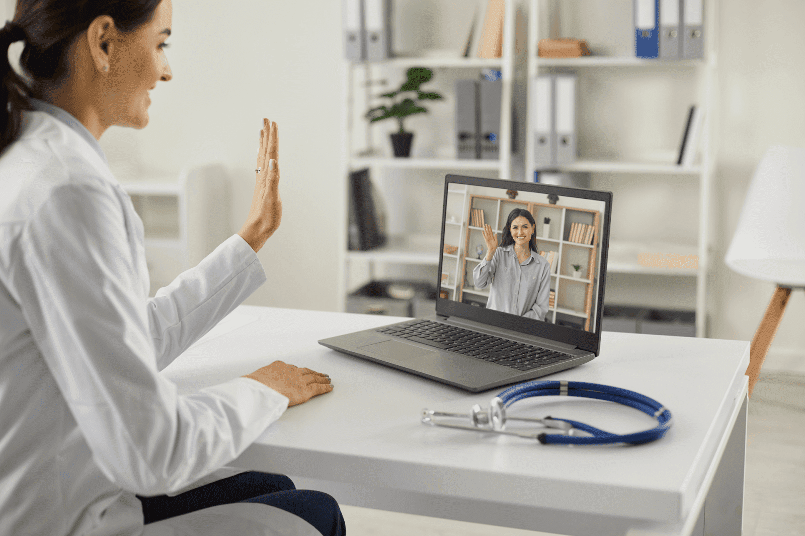 A female doctor waves while on a video call with a patient on her laptop