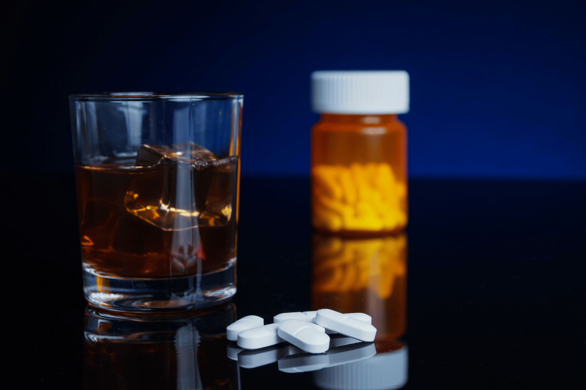 A Glass of alcohol beside white pills and a medicine bottle on a dark surface.