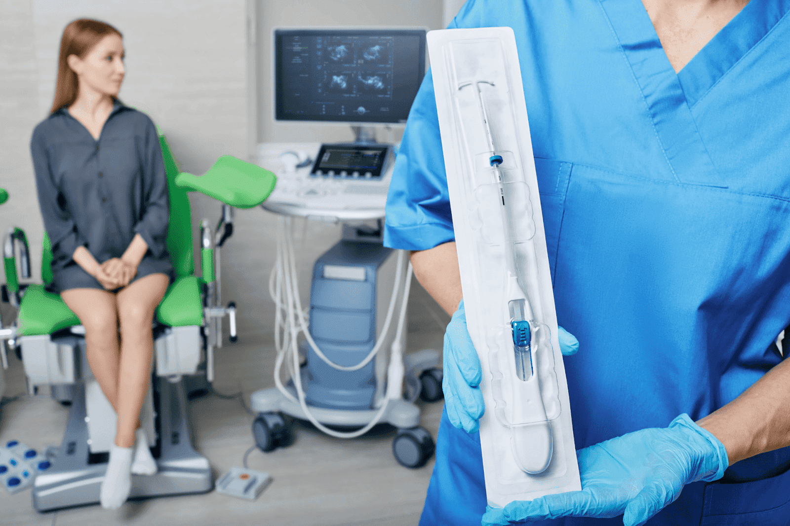 A healthcare provider holding a packaged IUD device while a patient waits in a gynecology exam room.