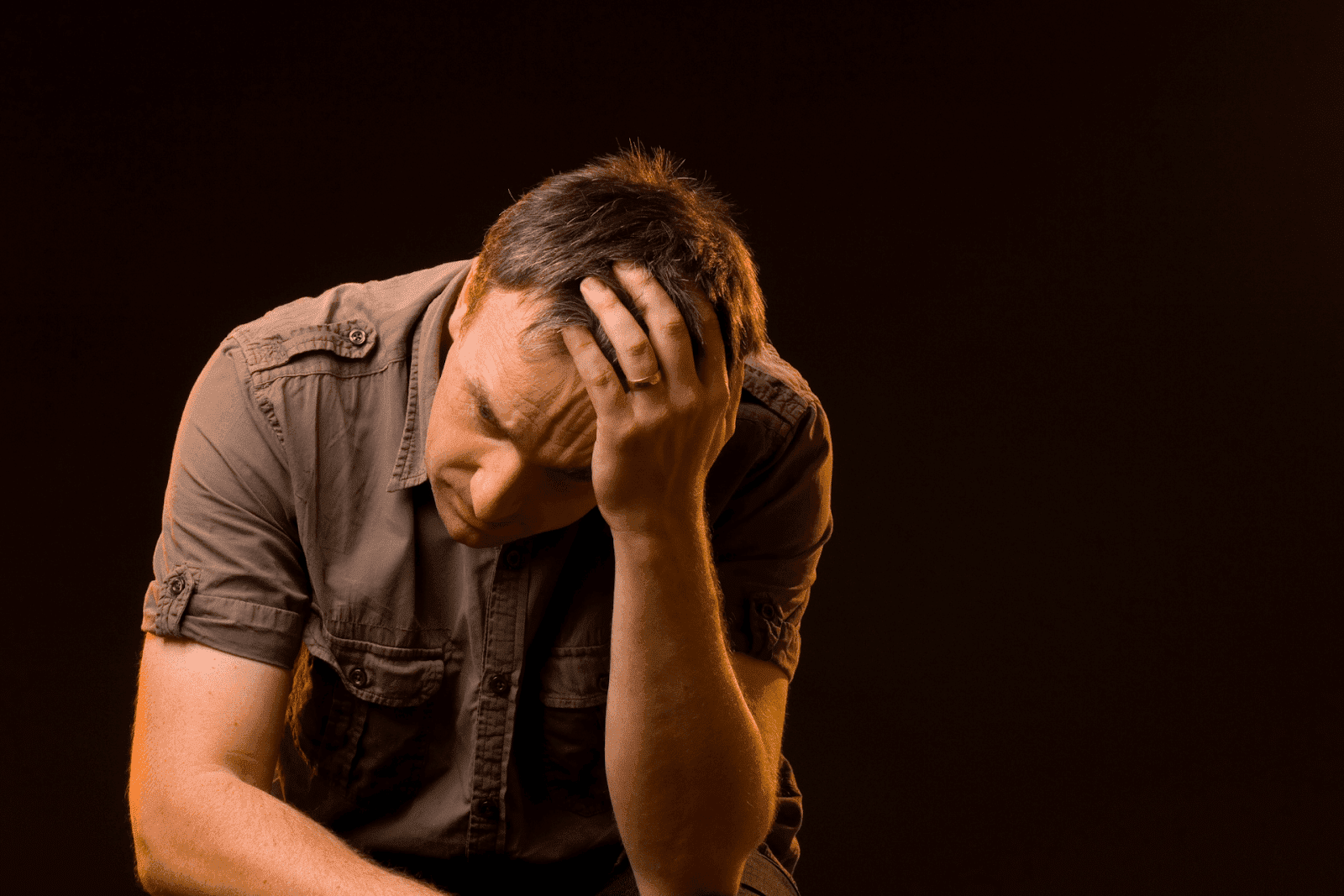 A man is sitting in a dark setting, hunched forward with his hand covering part of his face.