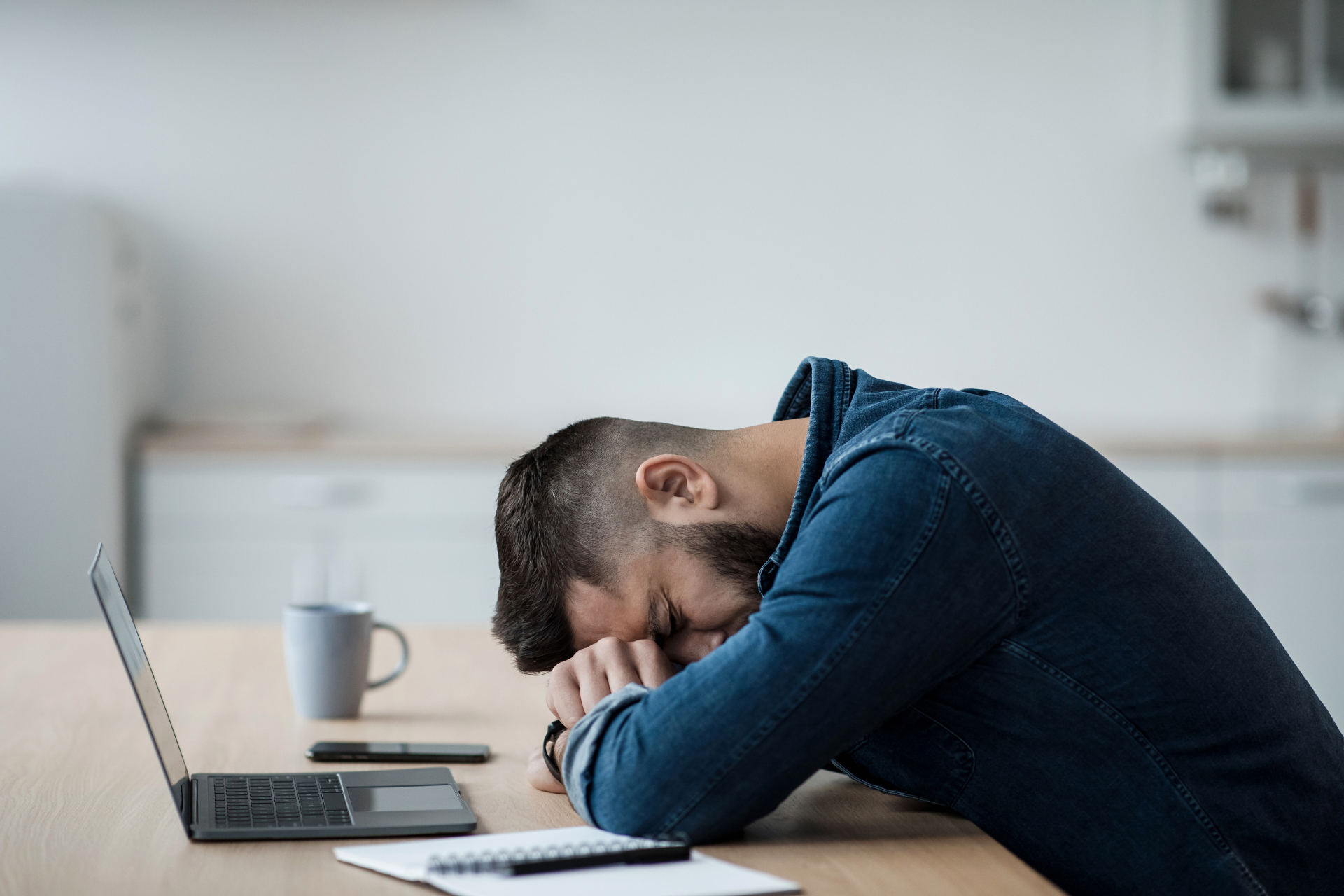 A man rests his head on his arms at a desk beside an open laptop, appearing tired or stressed