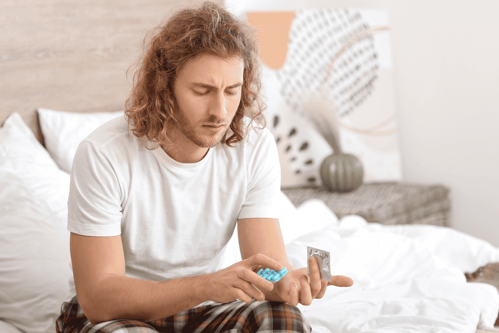A man sitting on a bed holding a pack of blue pills and a foil packet, looking down thoughtfully.