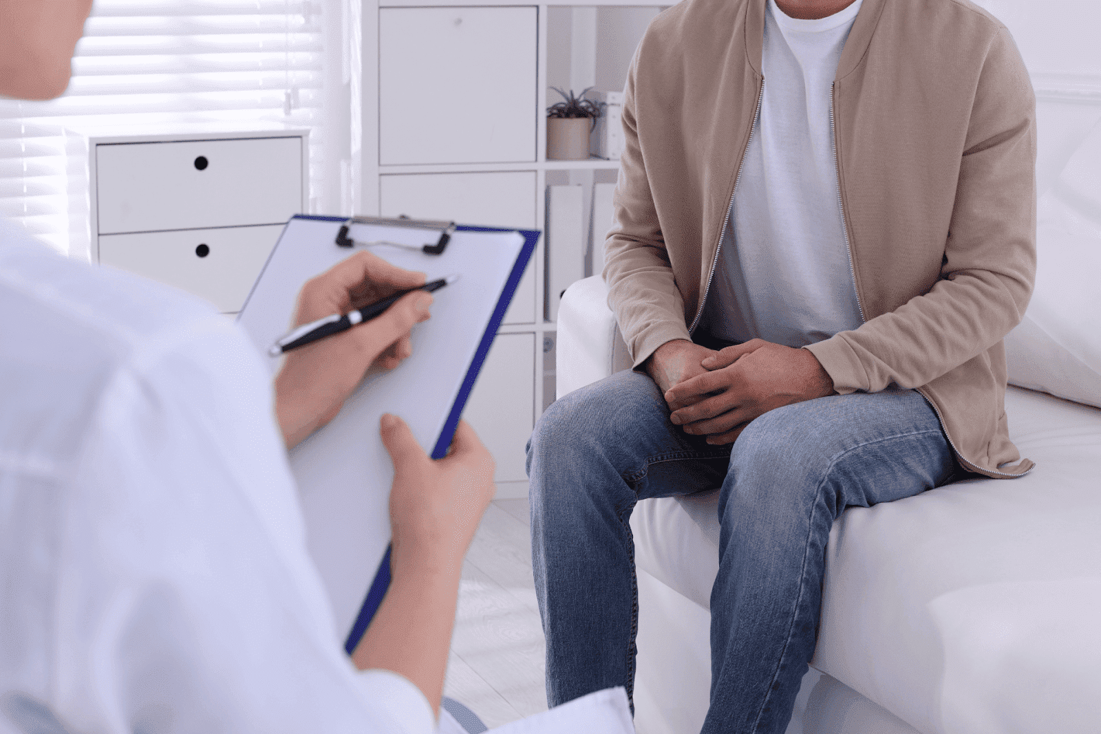 A man sitting on a couch during a consultation with a healthcare professional holding a clipboard.