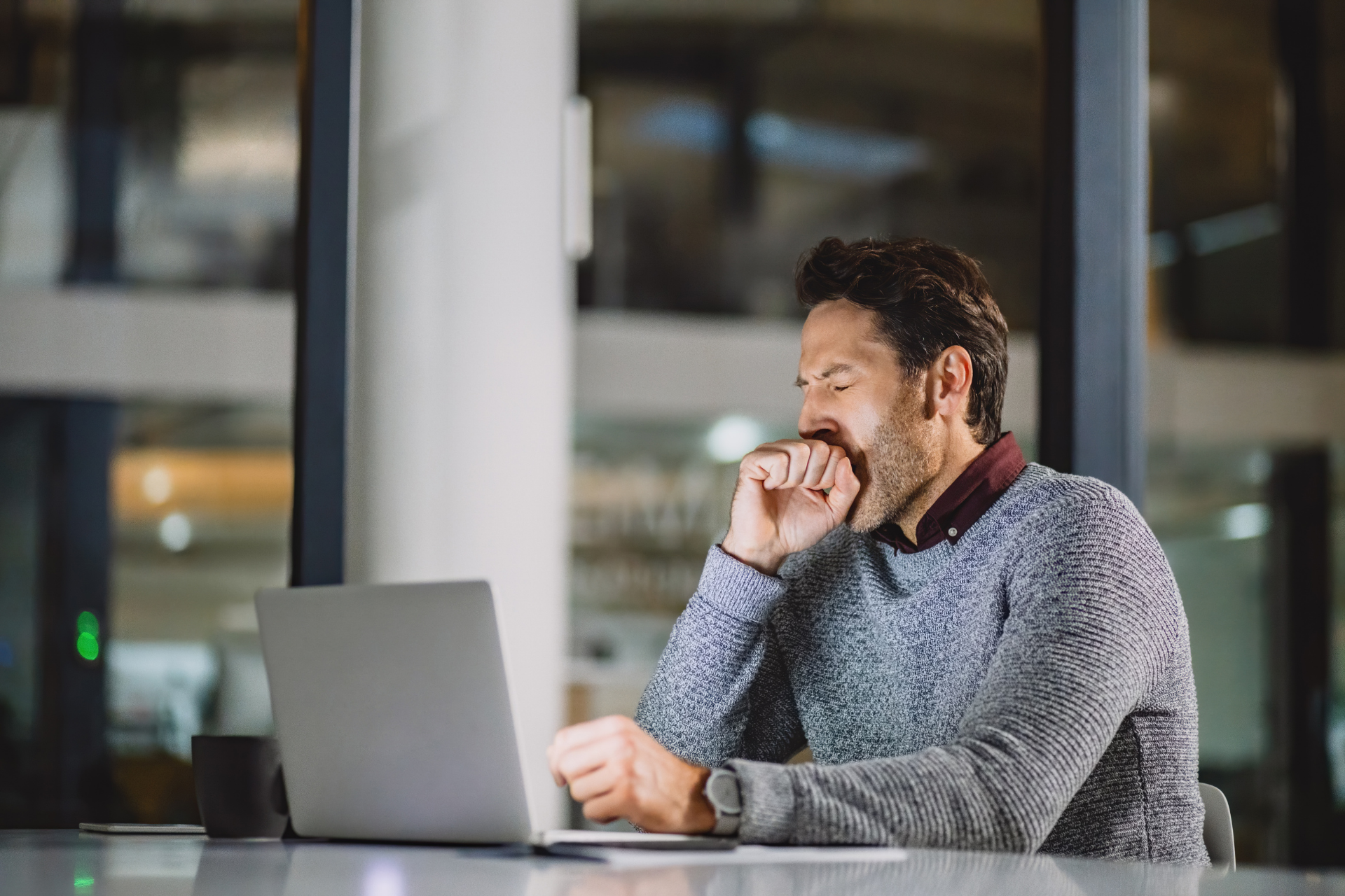 A man yawns while working on a laptop at a desk