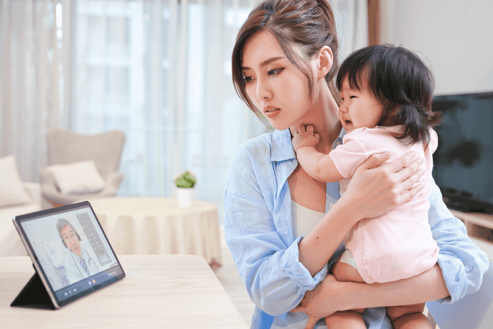 A mother holds her baby while having a video consultation with a doctor on a tablet