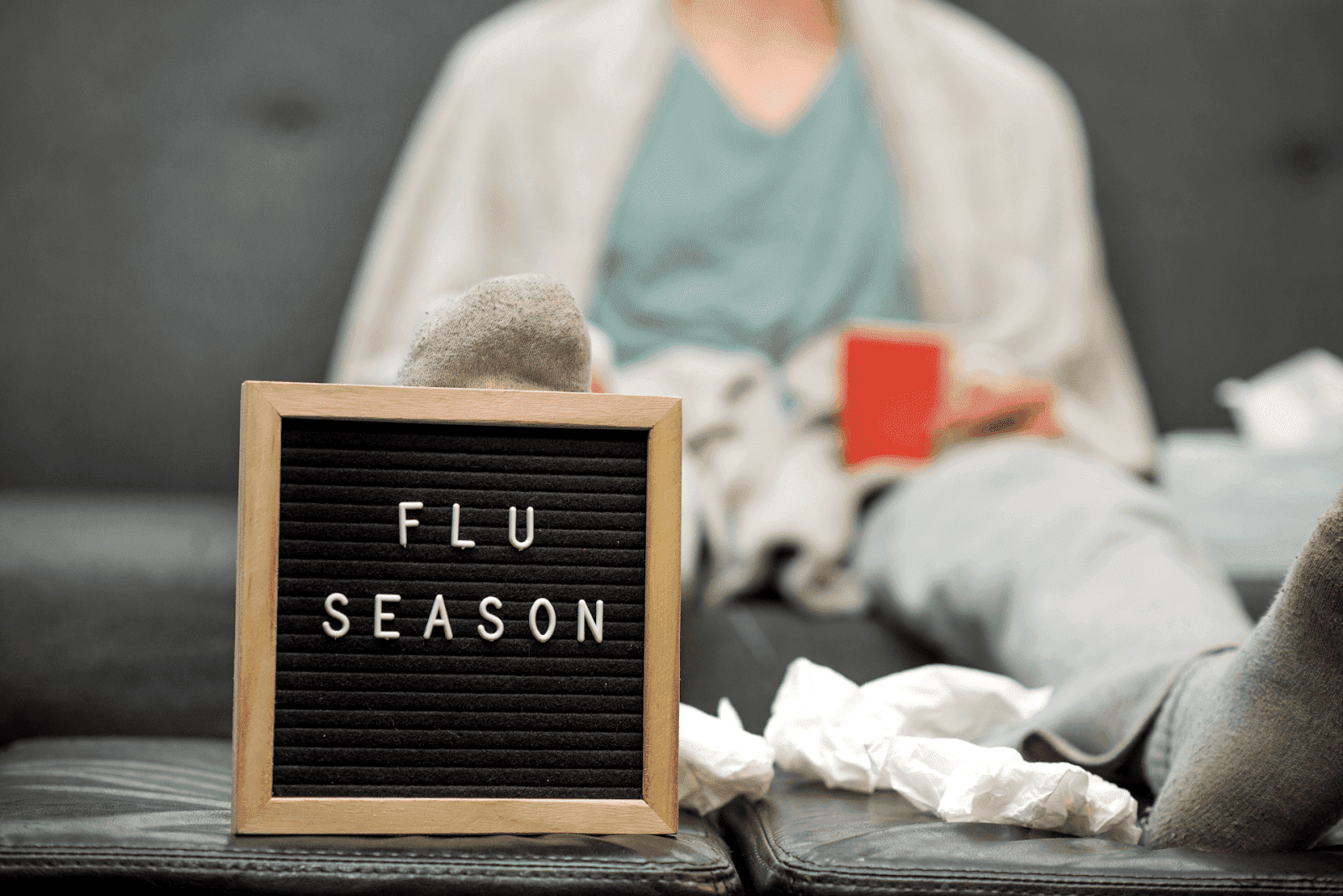 A person lying on a couch surrounded by tissues with a small sign in front that reads Flu Season.