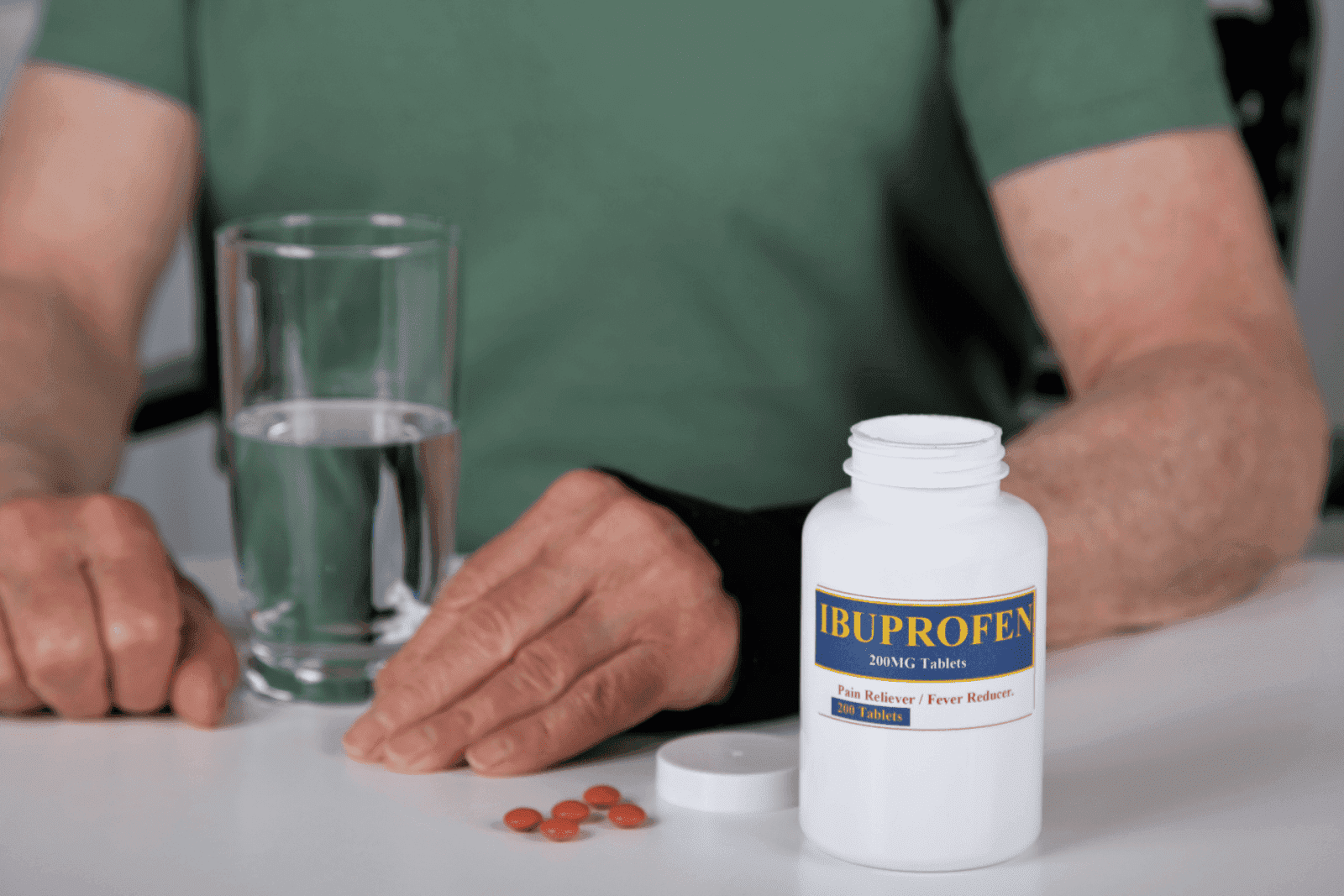 A person sitting at a table with an open bottle of ibuprofen, several tablets, and a glass of water