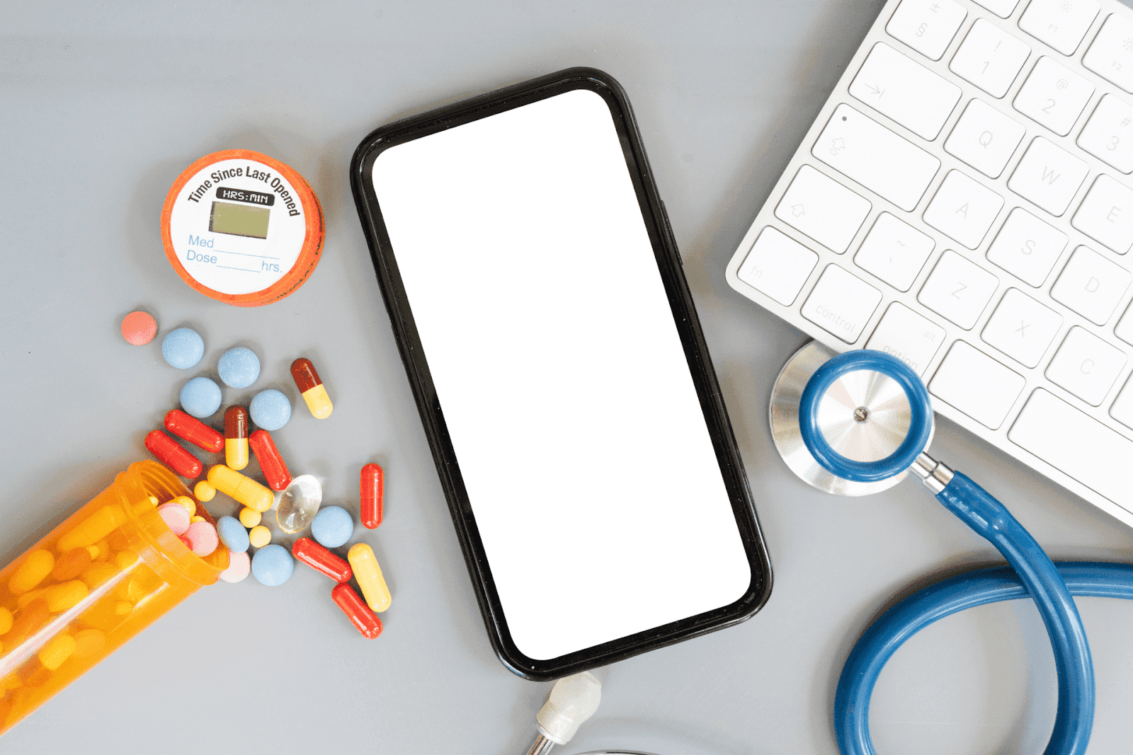 A smartphone on a desk surrounded by colorful pills, a stethoscope, a prescription bottle, and a keyboard