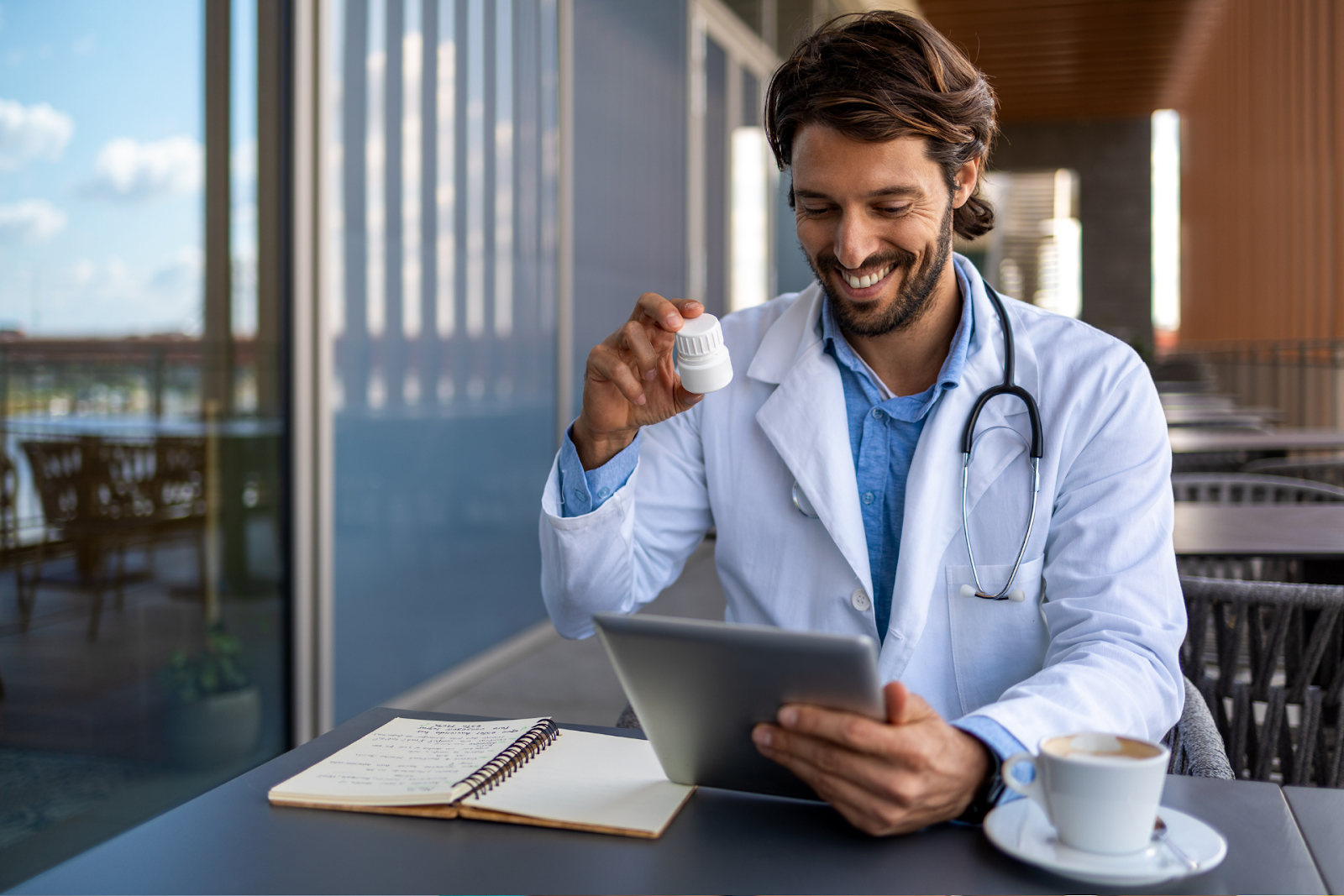 A smiling doctor in a white coat holding a pill bottle while looking at a tablet, with a notebook and a coffee cup on the table