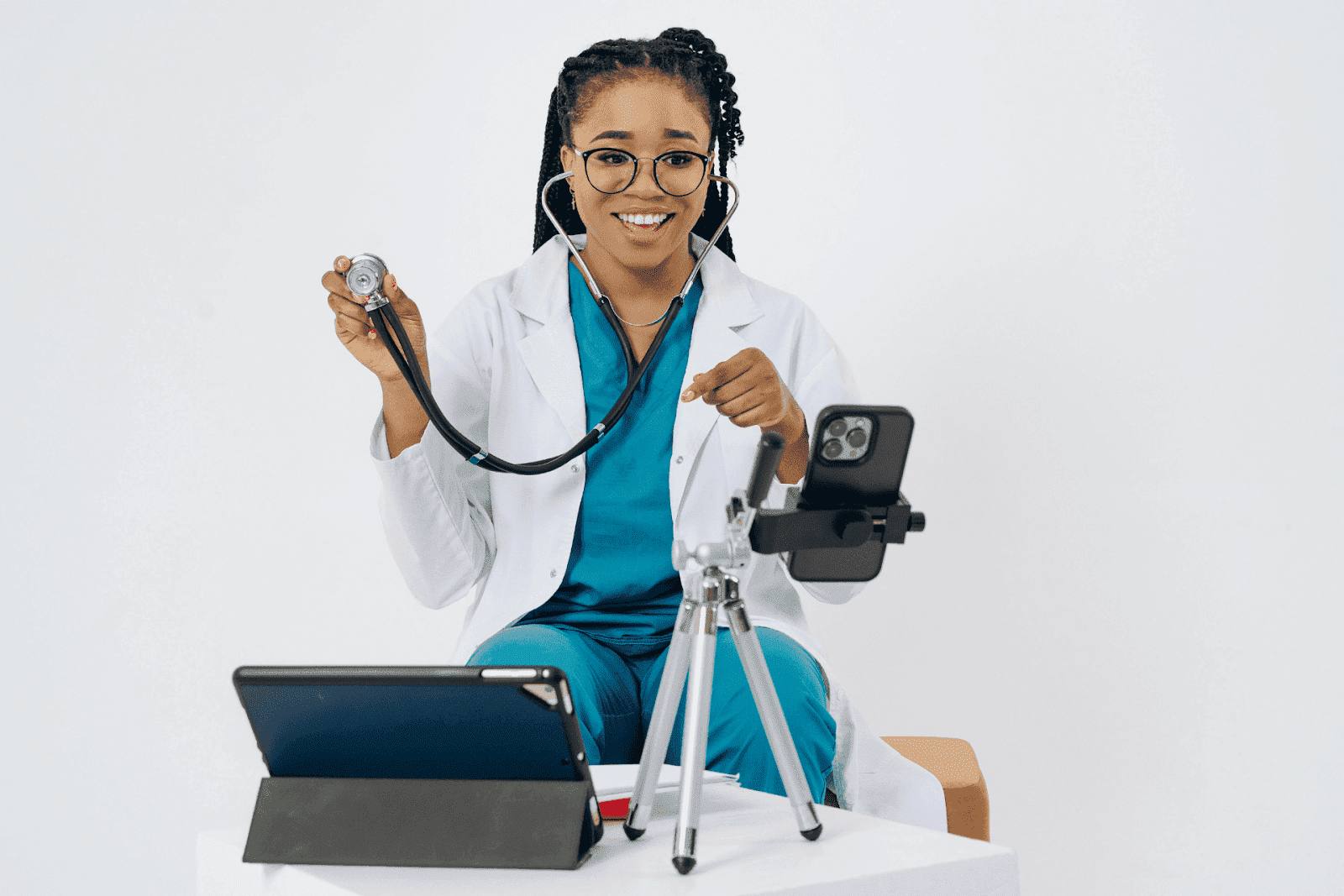 A smiling doctor in a white coat holds a stethoscope while sitting in front of a phone and tablet for a telehealth session