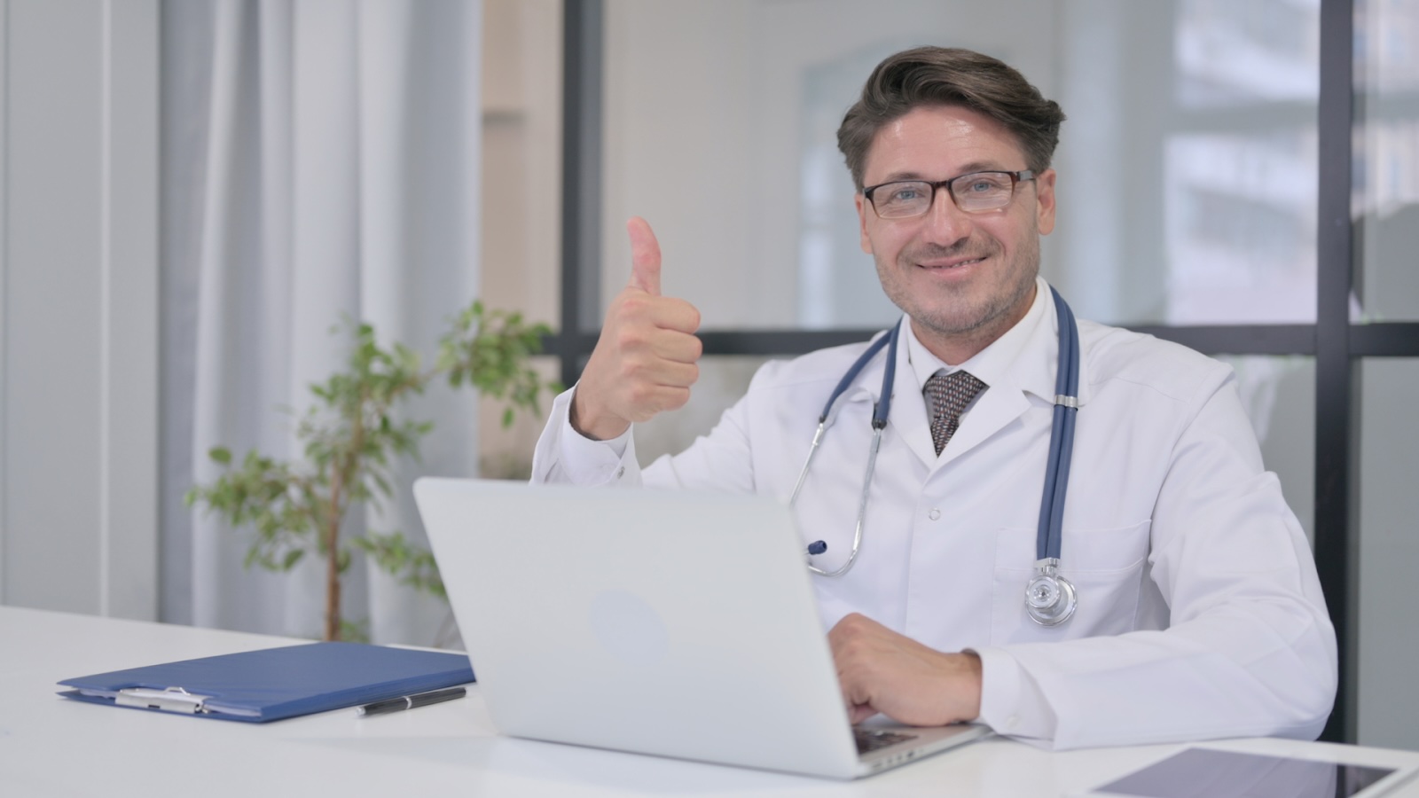 A smiling male doctor in a white coat giving a thumbs-up while sitting at a desk with a laptop
