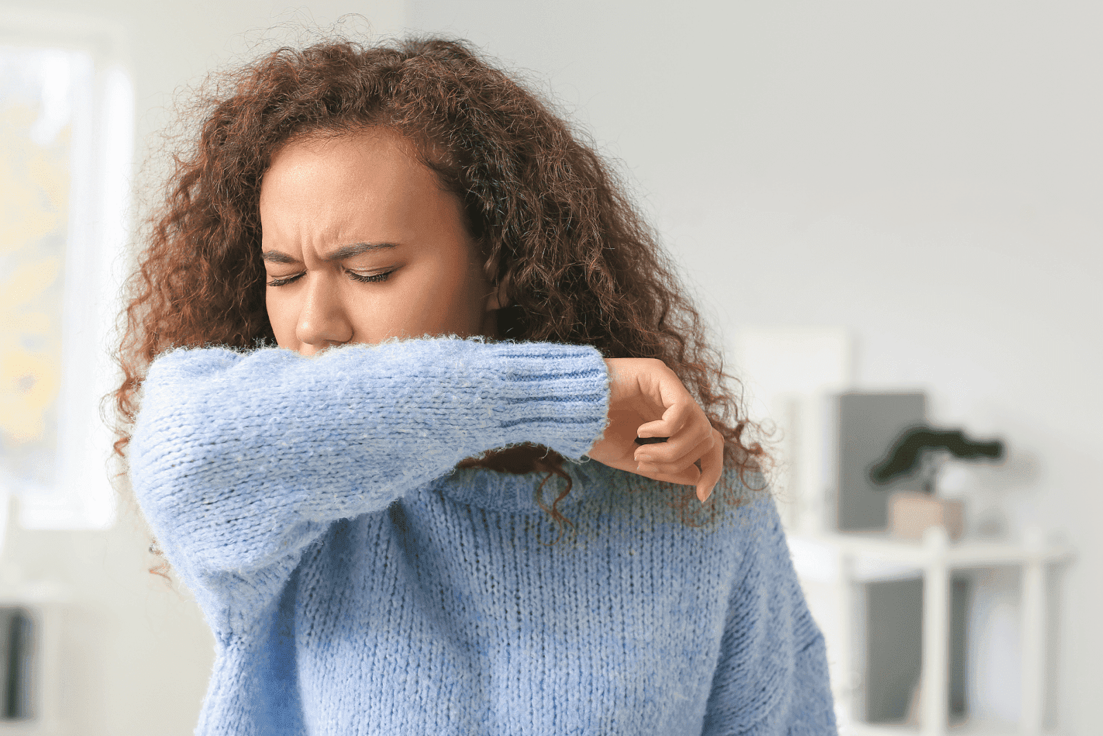 A woman coughing into her elbow while wearing a light blue sweater.