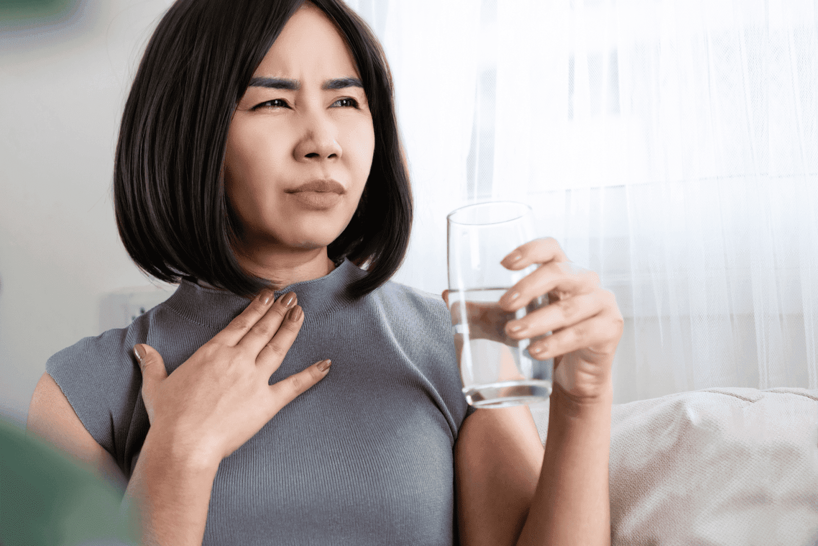A woman holding a glass of water while touching her chest and appearing uncomfortable
