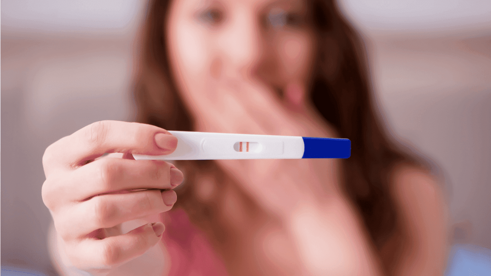 A woman shows a positive pregnancy test with two lines, covering her mouth in surprise.