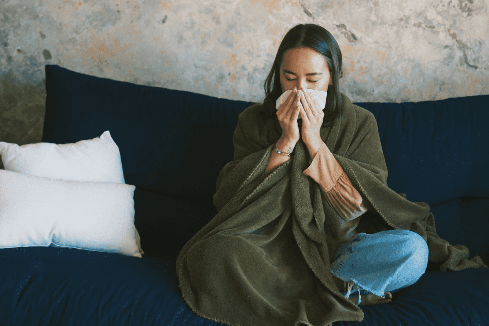 A woman sitting on a couch wrapped in a blanket, blowing her nose into a tissue.