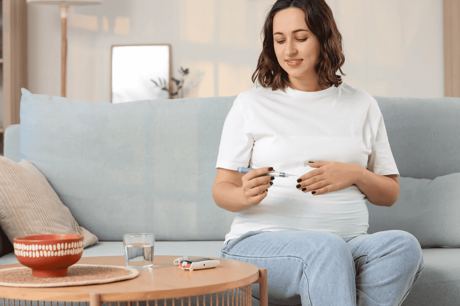 A woman sitting on a sofa prepares an insulin injection, holding an insulin pen against her abdomen, with a glucose meter and a glass of water on a small table nearby in a bright living room.