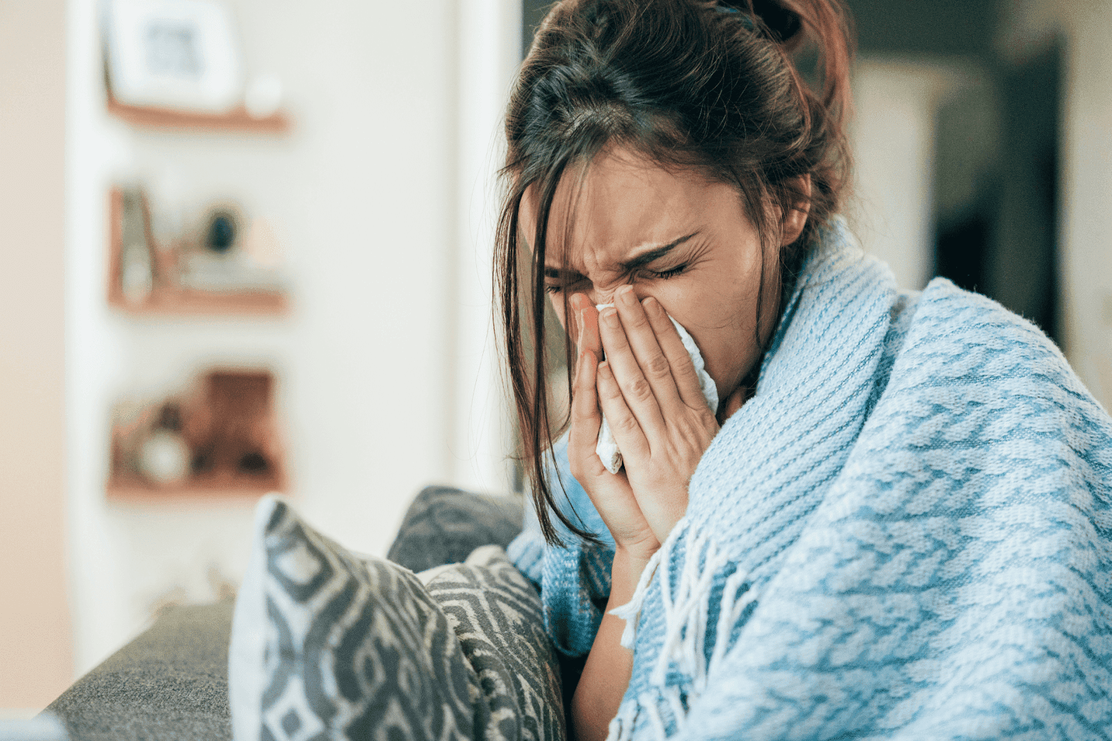 A woman sneezing into a tissue while wrapped in a blanket