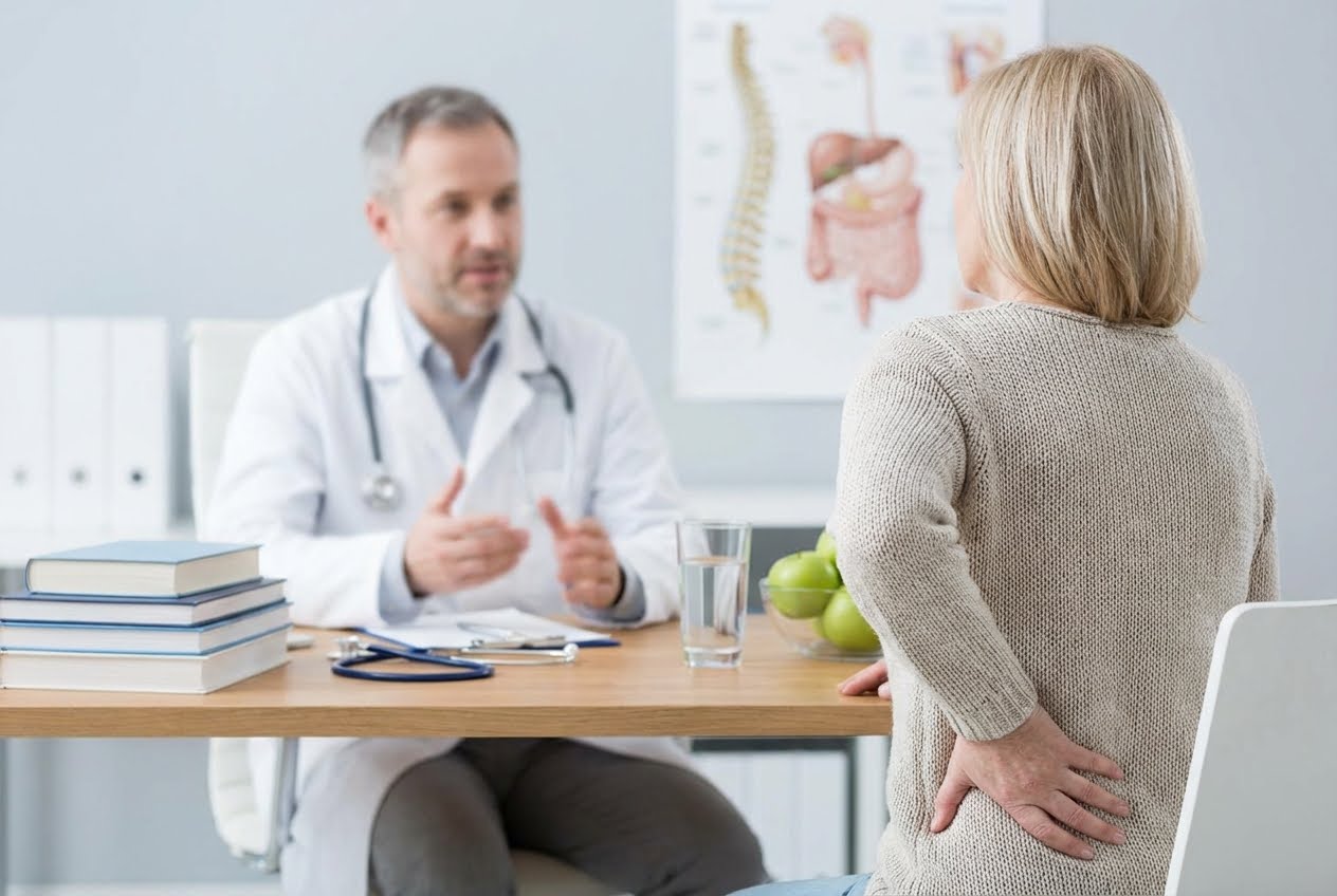 A woman with her back to the camera holds her lower back while a doctor sits across from her at a desk.
