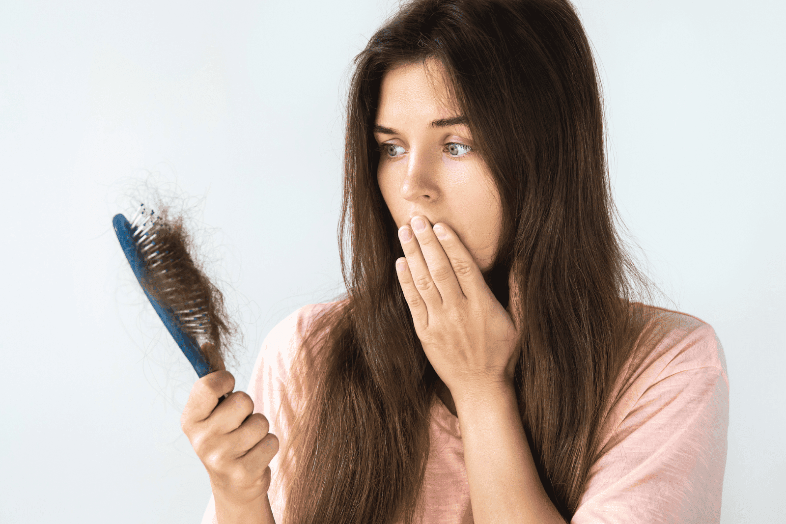 A woman with long brown hair looks worried as she holds a hairbrush filled with shed hair, covering her mouth with her other hand against a plain light background.