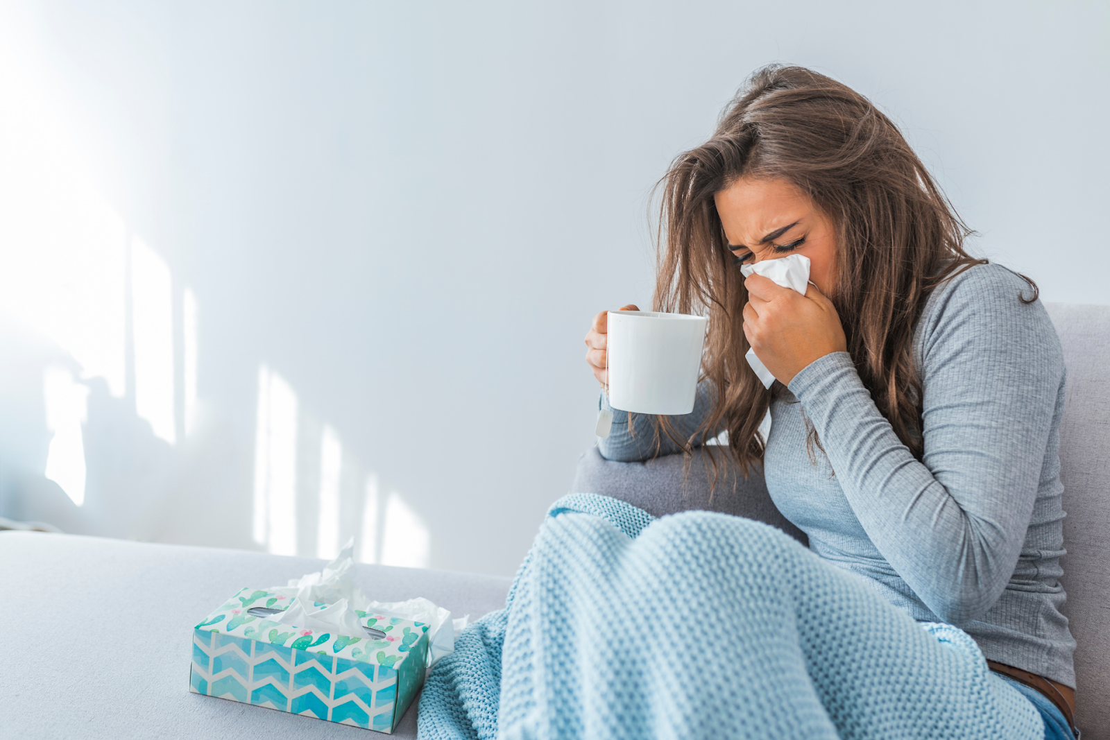 A woman wrapped in a blanket sitting on a couch, blowing her nose into a tissue while holding a mug.