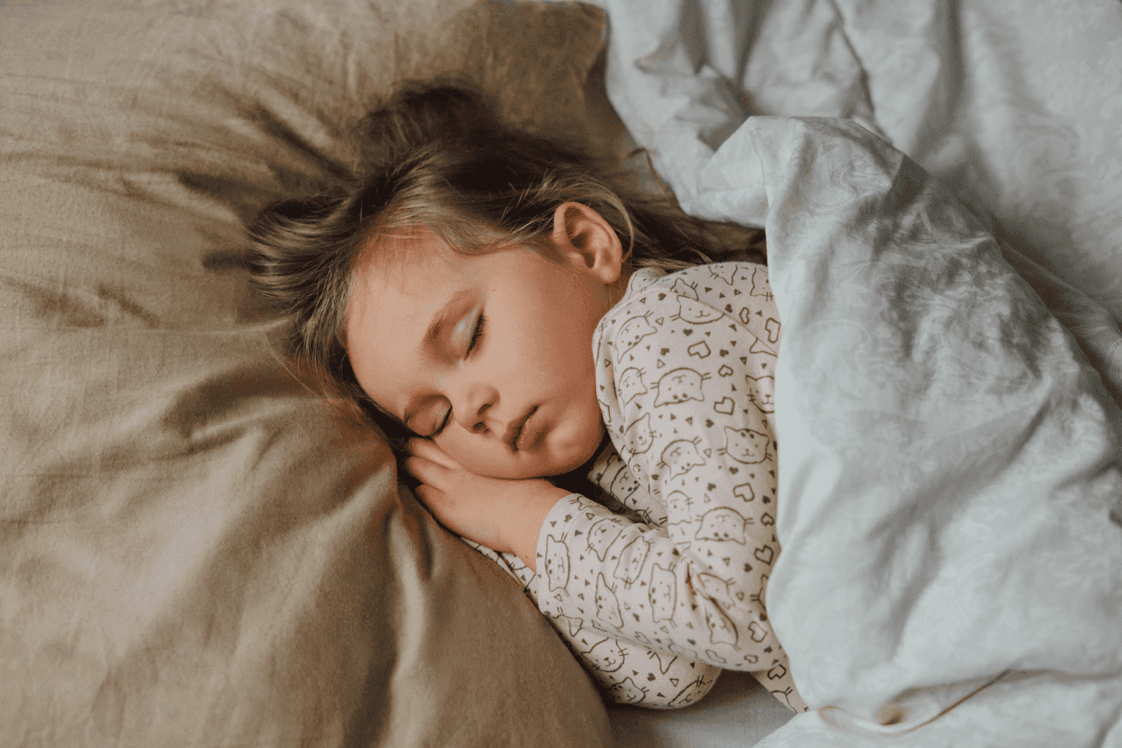 A young child is peacefully sleeping in bed, resting on a beige pillow and covered with a white blanket