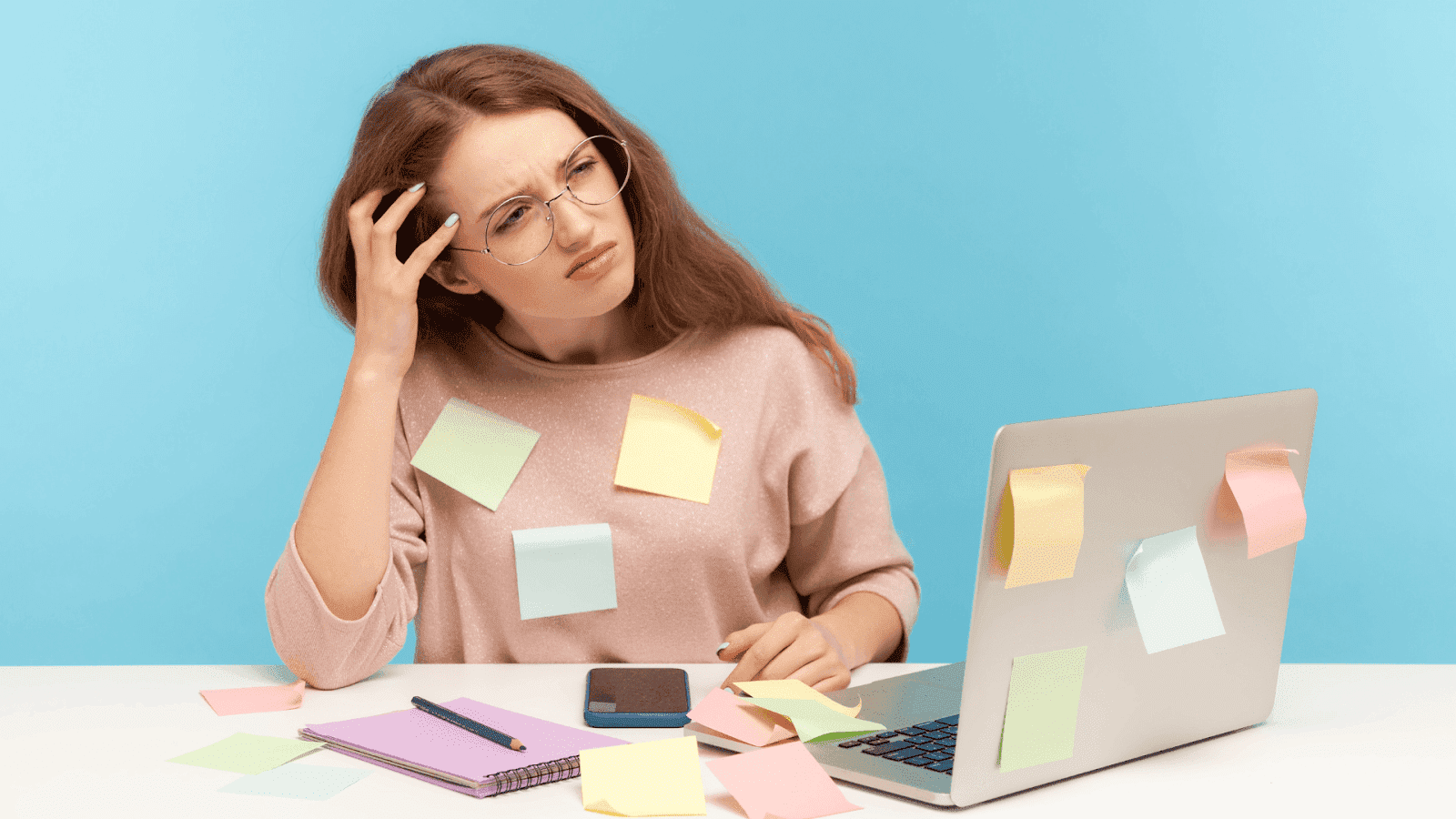 A young woman sits at a desk covered in sticky notes, looking confused while working on a laptop.