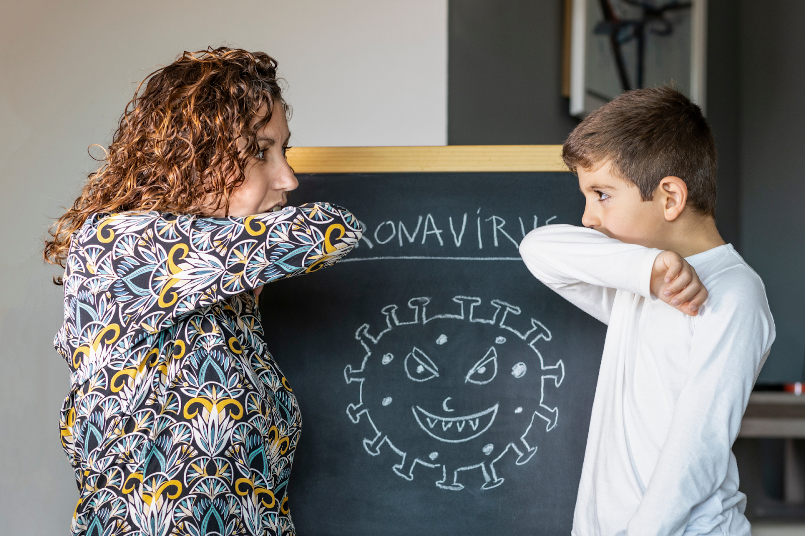 Adult and child demonstrating proper cough etiquette in front of a coronavirus illustration on a chalkboard