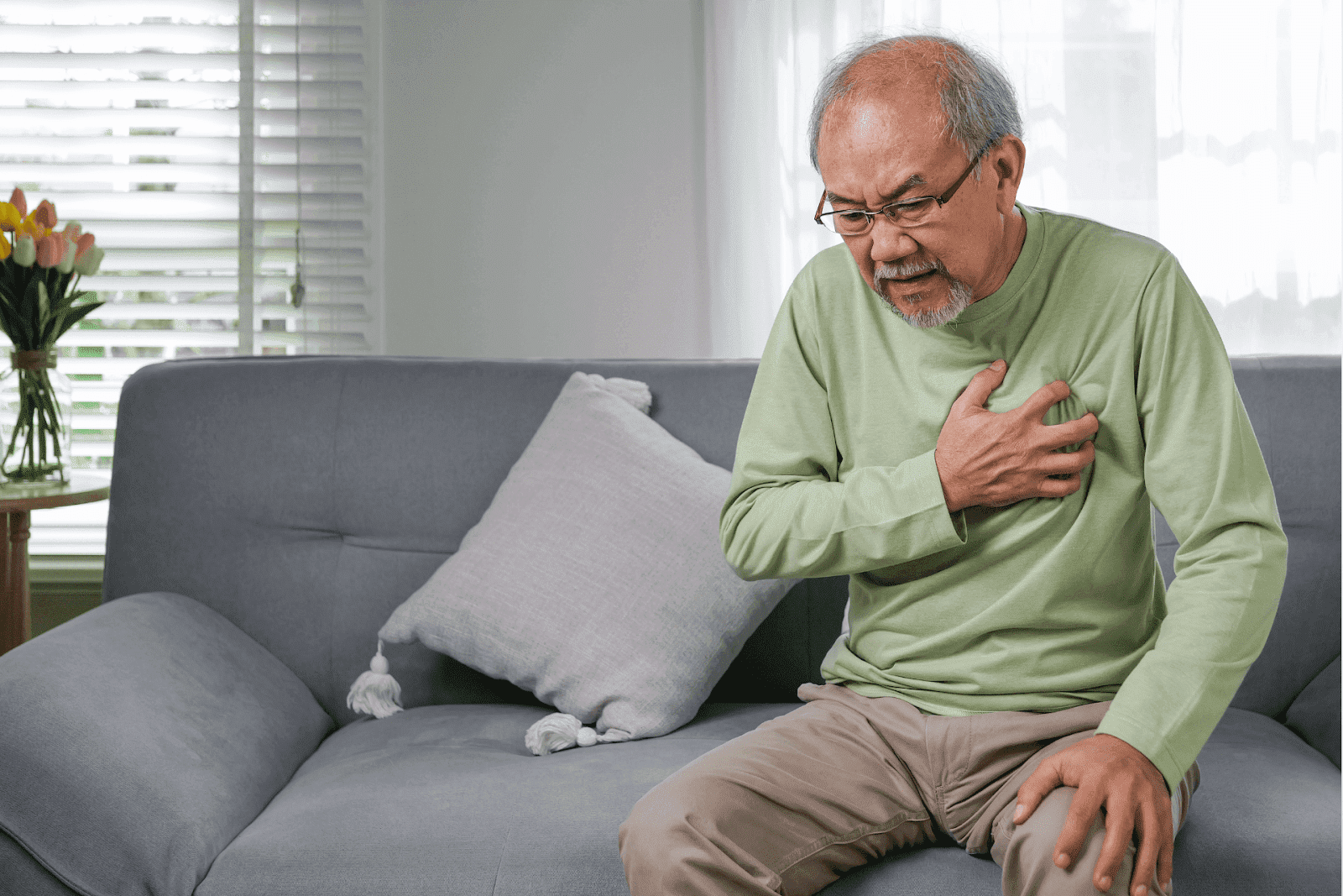 An elderly man is sitting on a sofa, holding his chest, suggesting chest pain or discomfort