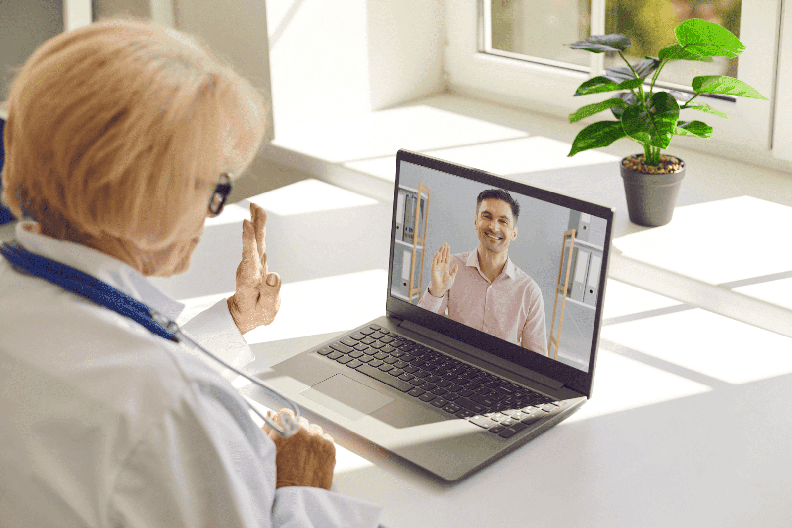 An older female doctor waves during a telehealth session with a smiling male patient on a laptop screen