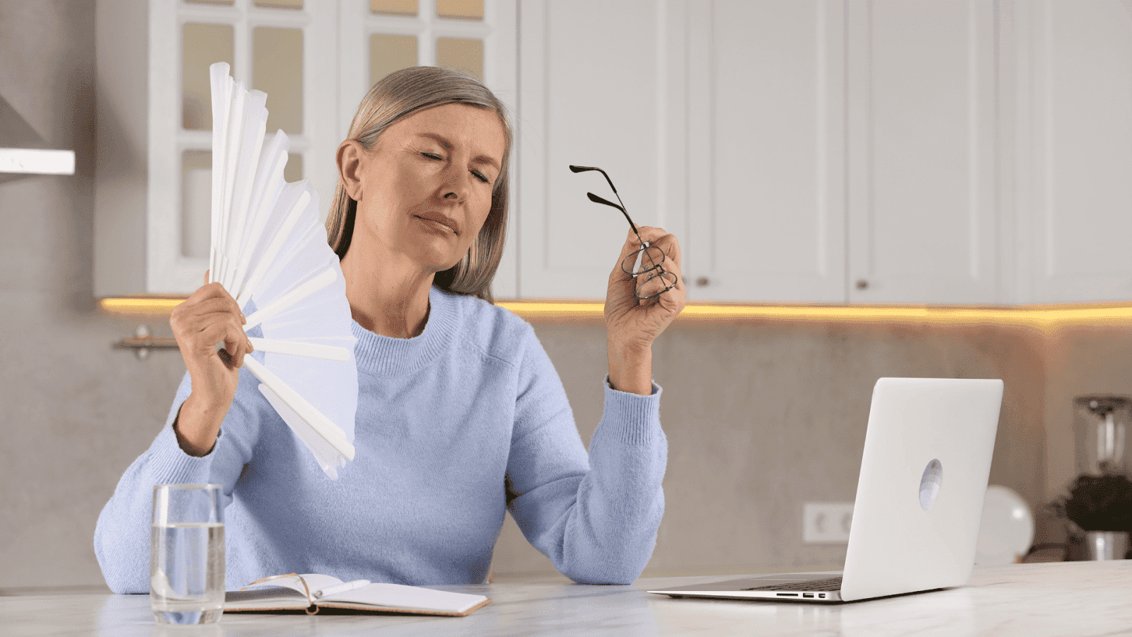 An older woman in a kitchen fanning herself while holding her glasses, suggesting a hot flash or menopausal symptom