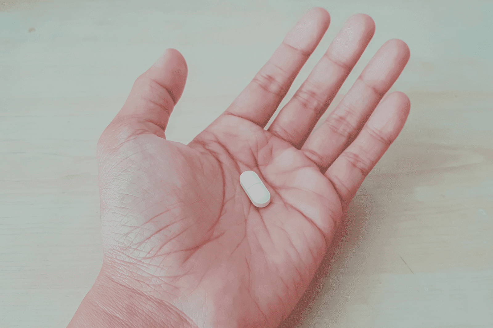 An open palm holding a single white oblong pill against a light wooden surface, representing medication or supplement use.