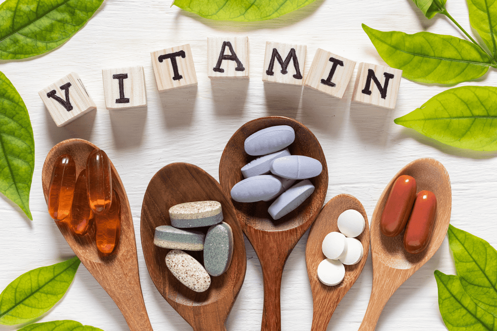 Assorted vitamin tablets and capsules arranged on wooden spoons with green leaves and wooden letter blocks spelling "VITAMIN".
