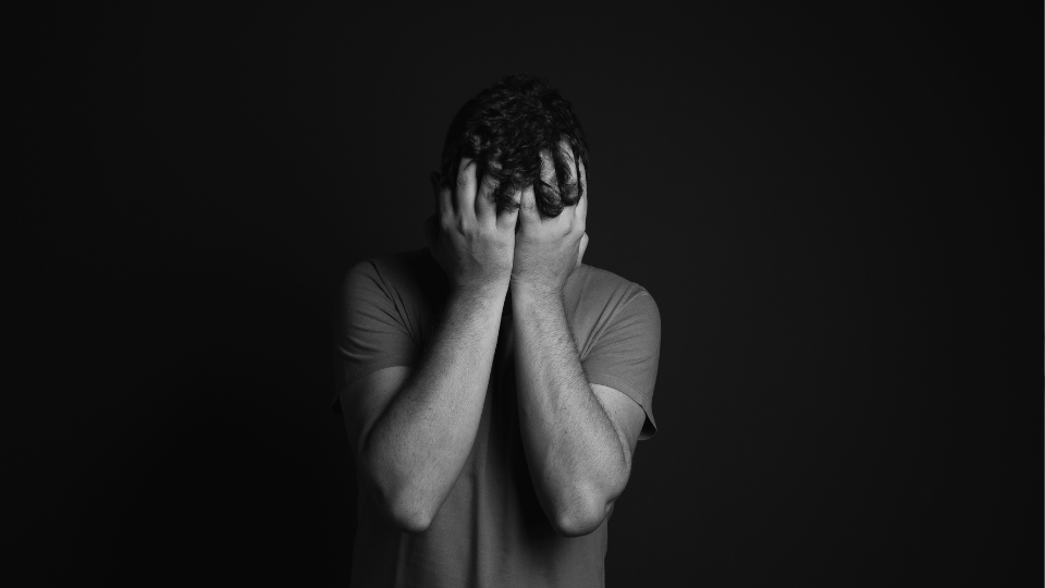 Man standing against a dark background with his face buried in his hands, expressing emotional distress.