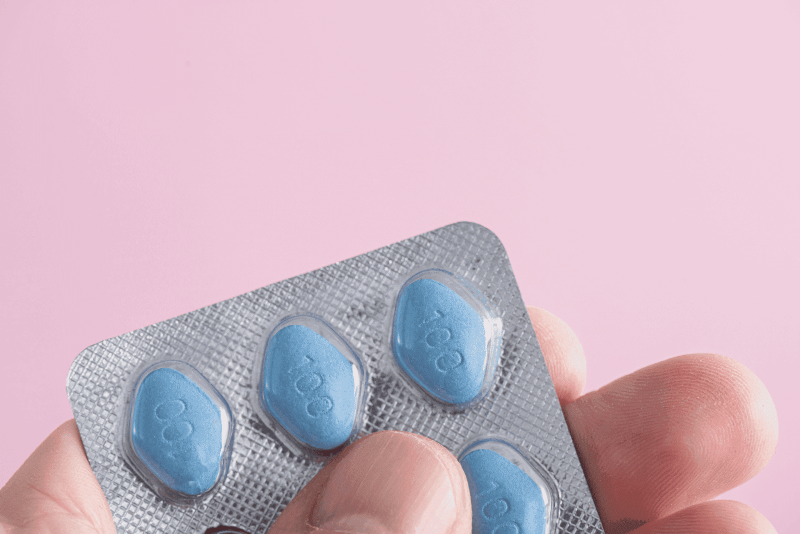 A hand holds a blister pack containing four blue, diamond-shaped pills marked with “100,” set against a soft pink background.