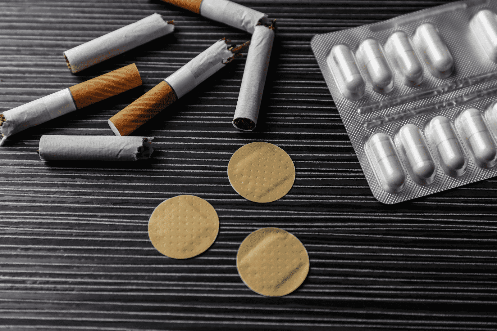 broken cigarettes, nicotine patches, and a blister pack of capsules arranged on a dark surface, symbolizing smoking cessation aids.
