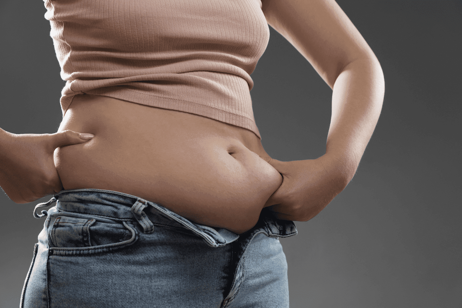 Close-up of a woman pinching her belly fat while wearing jeans and a crop top.