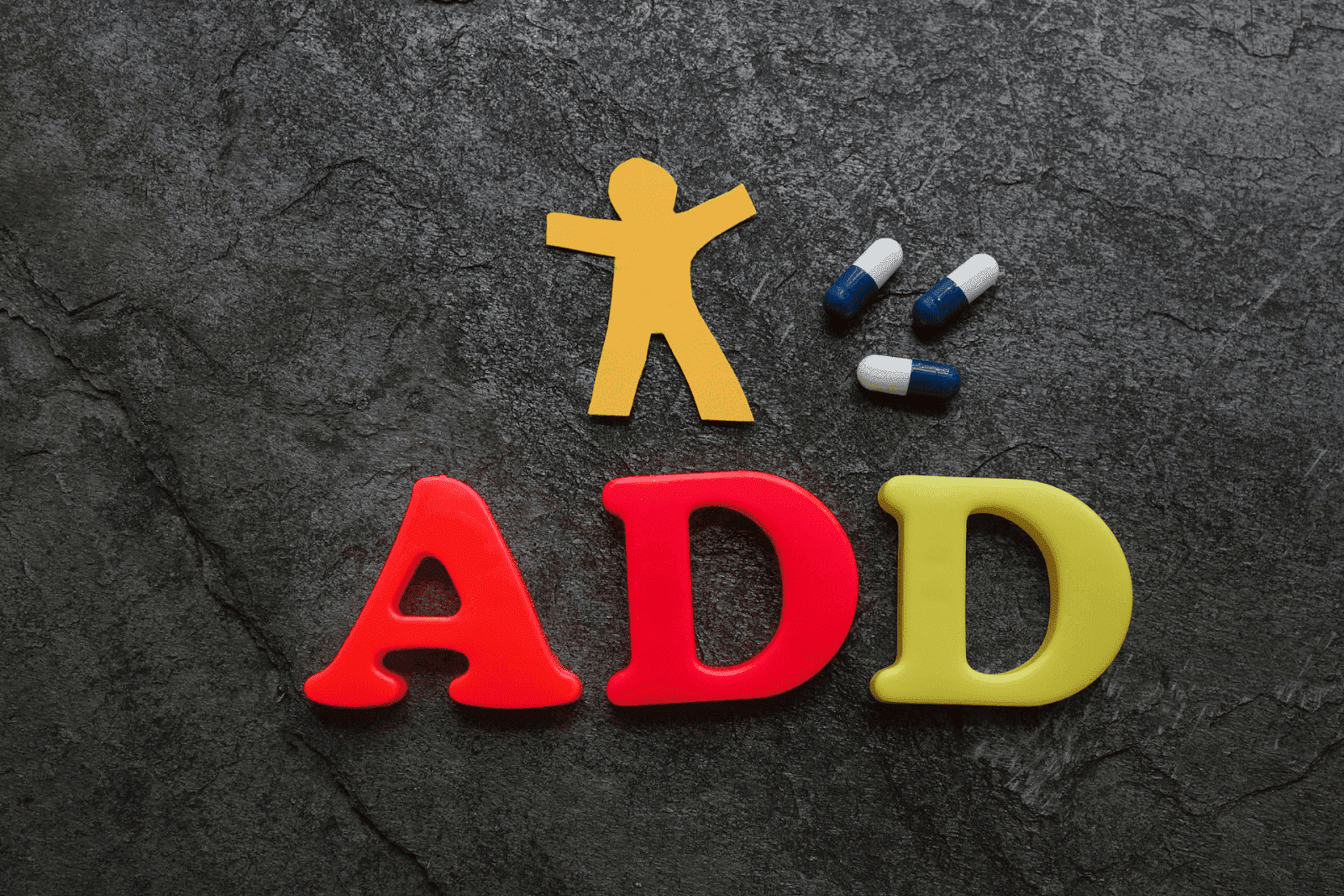 Colored letters spelling "ADD" with paper cutout figure and capsules on dark background.