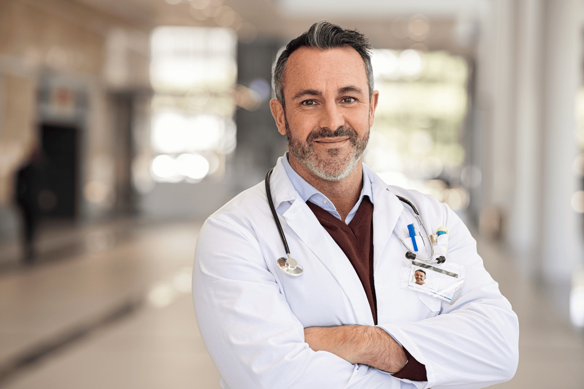 Confident male doctor in a white coat with arms crossed in a hospital hallway.