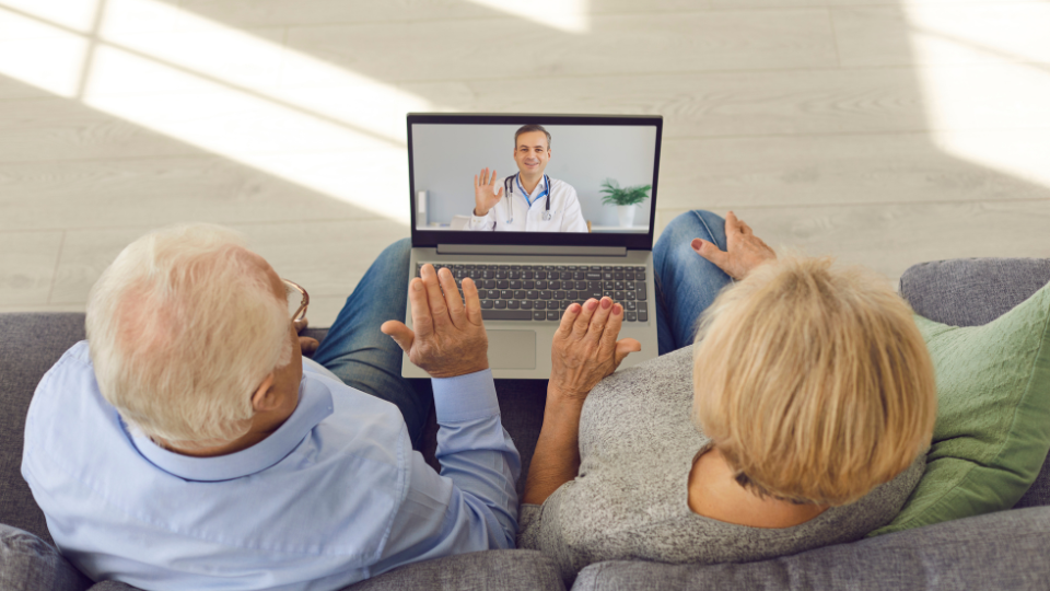 Elderly couple waving into laptop on telehealth video call with doctor waving back
