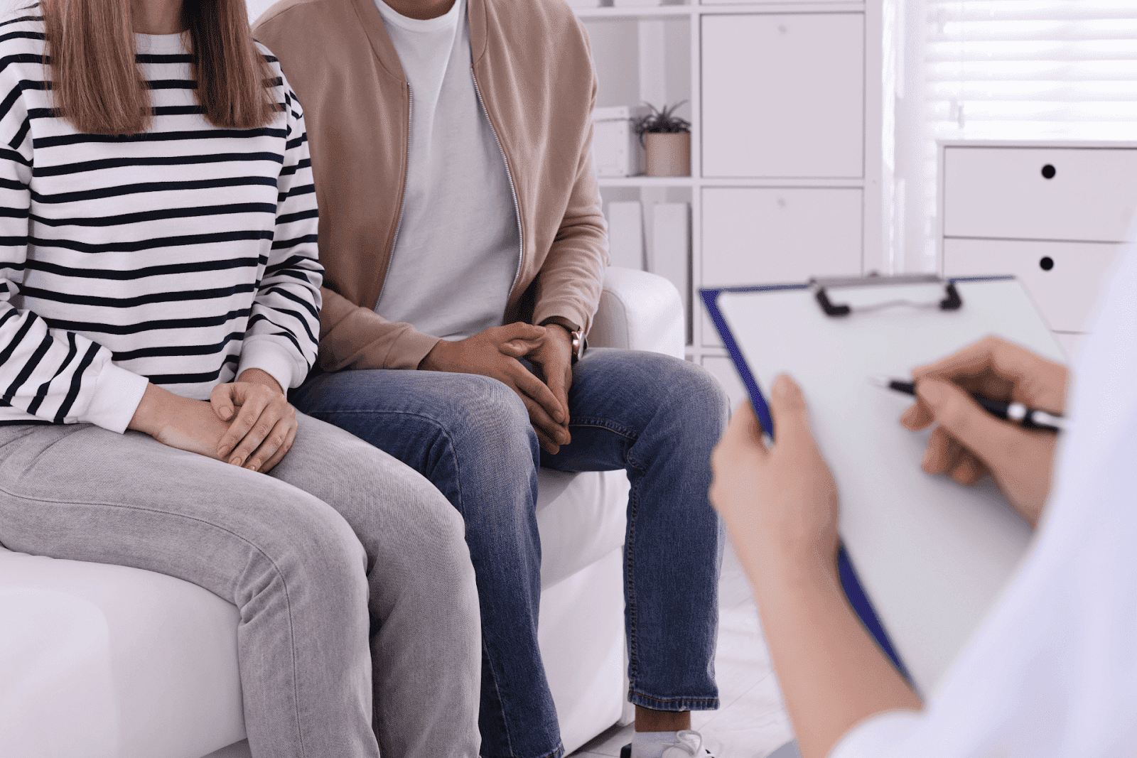Couple sitting on a couch during a consultation, facing a healthcare professional holding a clipboard.